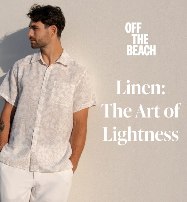 A man leaning against a wall, wearing a floral-patterned linen shirt and white linen pants. 